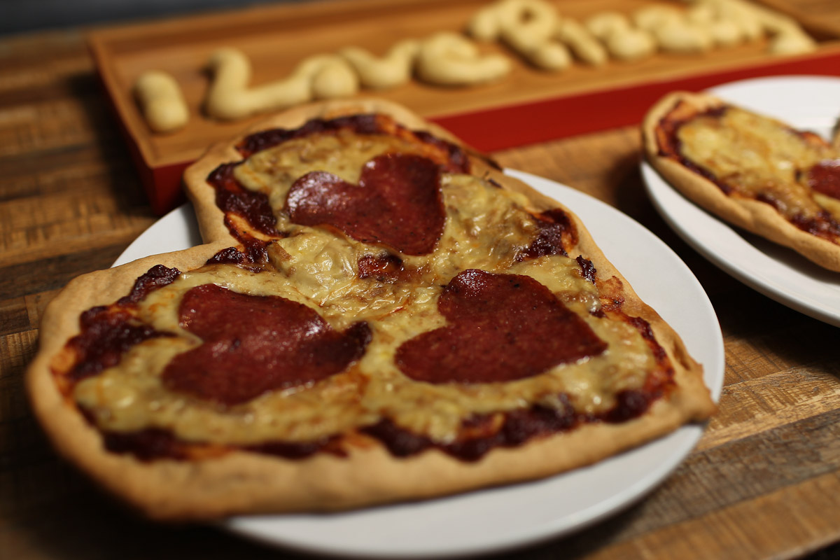 A glistening heart shaped pizza with heart shaped ingredients is viewed from a side angle. You can see there is a second pizza in the background as well as bread that spells 