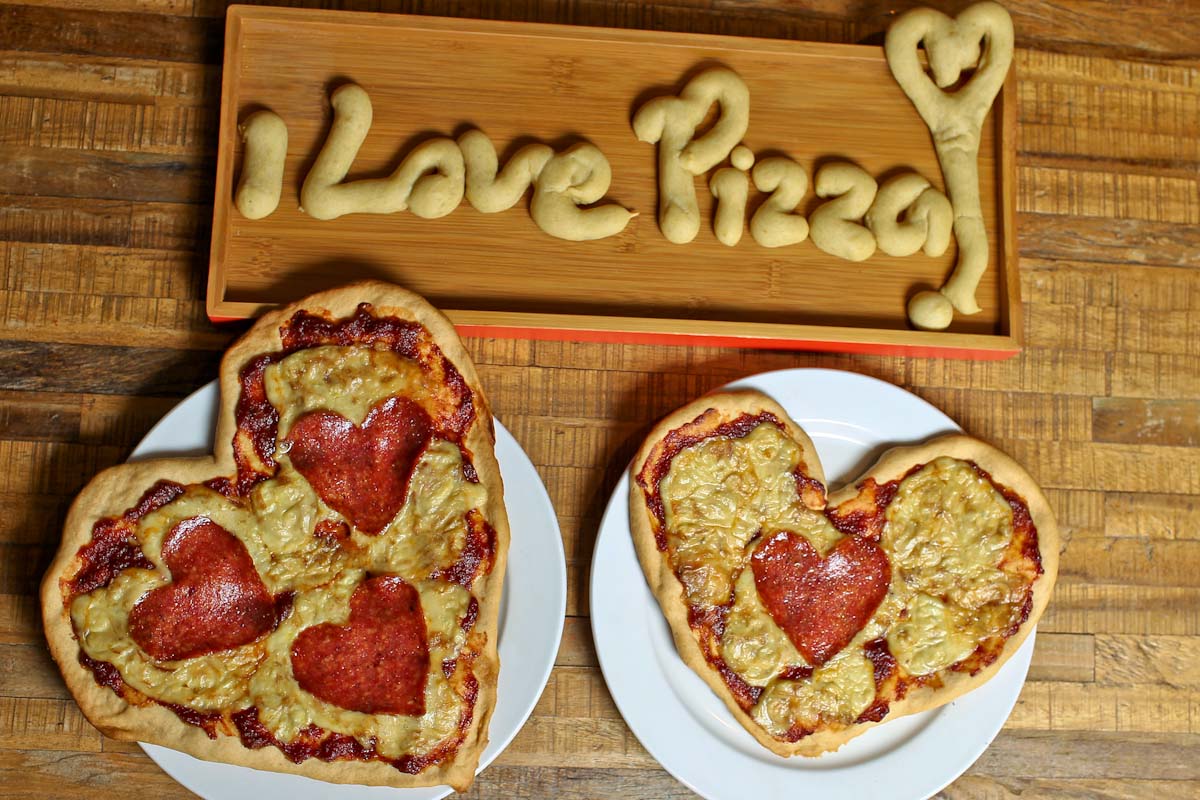 Two heart shaped gluten free pizzas are on a table. They have heart shaped ingredients on them. There is bread that says 