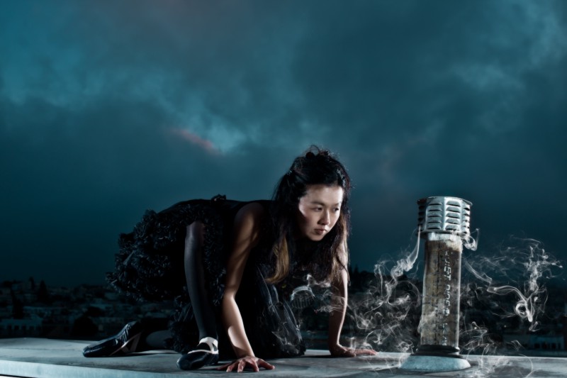 A ballerina crouches with intent on a rooftop. Her face is near the smoke drifting out of a pipe venting in the roof top.