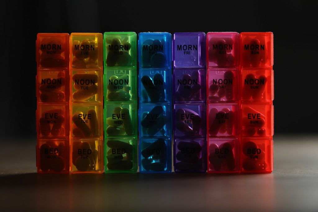 Photo of a pill organizer set. Each day is a different colour. Monday is red, Tuesday is yellow, Wednesday is green, Thursday is blue, Friday is purple, Saturday is pink, Sunday is orange. There are four slots for each day. Morning, Noon, Evening and Bedtime. 